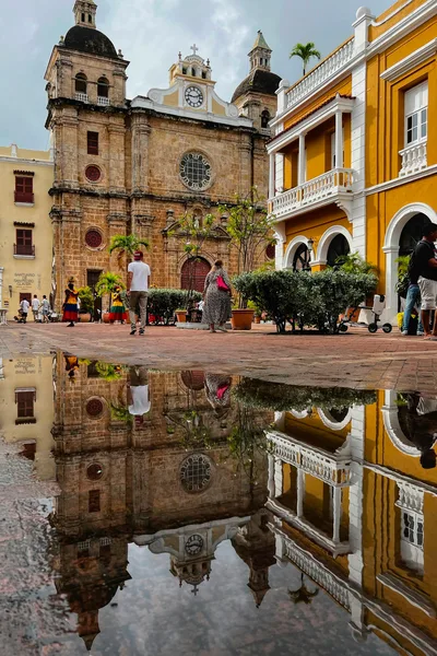 A bustling square in Cartagena, Colombia, featuring a historic church reflected in a puddle, capturing the vibrant urban scene.
