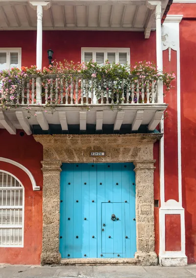 Charming colonial facade with vibrant red walls and a bright blue door, adorned by a flower-topped balcony.