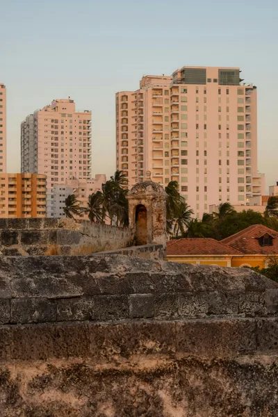 View of modern high-rise buildings behind an ancient fort wall at sunset.